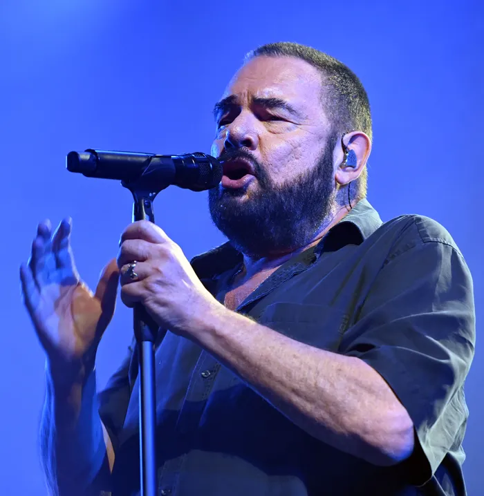 Ein bärtiger Mann singt in ein Mikrofon vor einem blau beleuchteten Hintergrund.