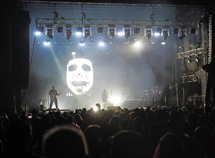 Die Band Alphaville tritt auf einer Bühne mit einer großen, leuchtenden Maske im Hintergrund vor einer dichten Menschenmenge auf.