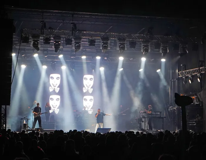 Alphaville performt auf einer hell erleuchteten Bühne mit Masken-Projektionen im Hintergrund vor einer Menschenmenge.