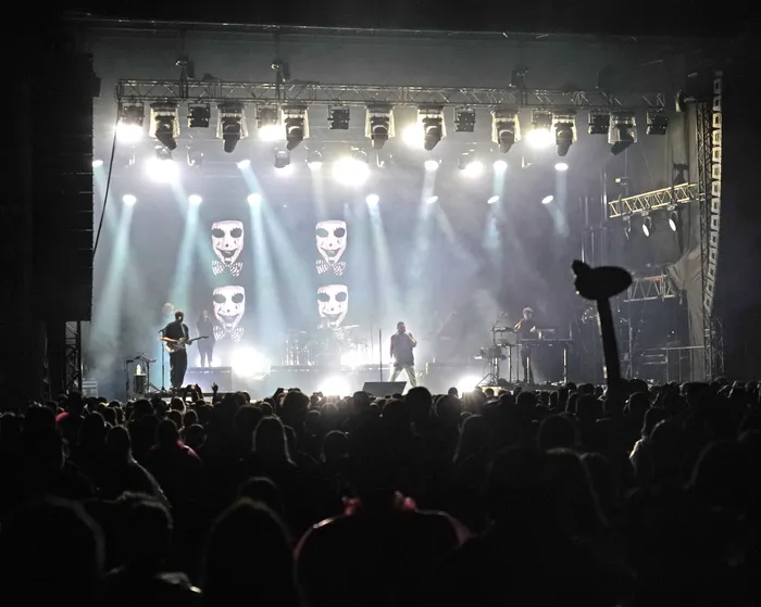 Die Band Alphaville tritt auf einer hell erleuchteten Bühne mit Scheinwerfern und grafischen Gesichtern vor einer großen Menschenmenge auf.