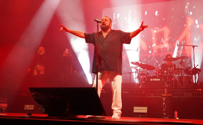 Der Sänger der Band Alphaville singt mit weit ausgebreiteten Armen in ein Mikrofon auf einer rot beleuchteten Bühne.
