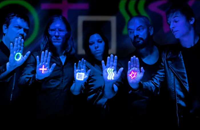 Fünf Mitglieder der Band Alphaville halten ihre Hände mit leuchtenden Neon-Symbolen wie einem Plus, einem Kreis und einem Stern in die Kamera.