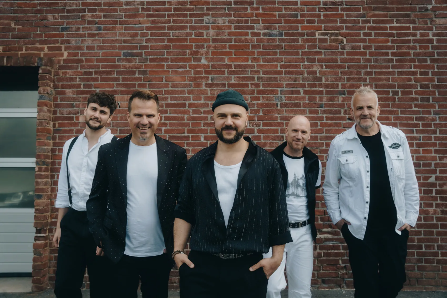 Fünf Männer stehen lächelnd vor einer Backsteinmauer. Sie tragen weiße Hemden und verschiedene Jacken oder Westen.