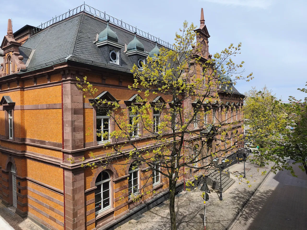 Zweigeschossiges Gebäude mit rotem Backstein und mehreren Fenstern, davor ein Baum mit frischem Grün und eine wenig befahrene Straße.