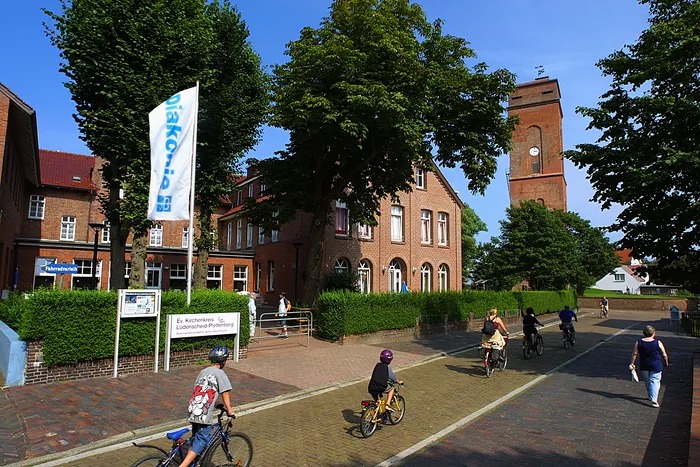 Backsteingebäude und ein hoher Turm hinter Bäumen, während Menschen auf Fahrrädern und zu Fuß eine gepflasterte Straße passieren.