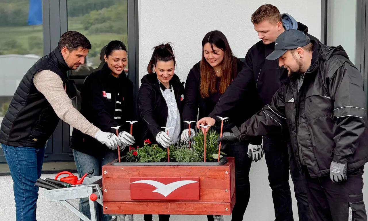 Sechs Mitarbeiter von Altrad plettac assco pflanzen gemeinsam Blumen in ein rotes Hochbeet mit weißem Logo.