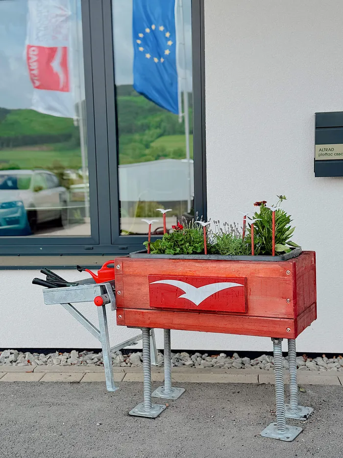 Rot gestrichenes Hochbeet aus Holz mit Blumen vor dem Eingang eines hellen Firmengebäudes.