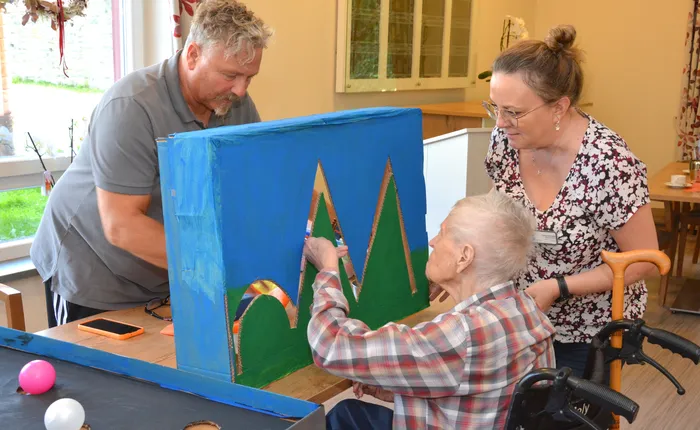 Ein älterer Mensch spielt mit einer blauen Box mit Bergmotiven, während ein Mann und eine Frau daneben stehen.