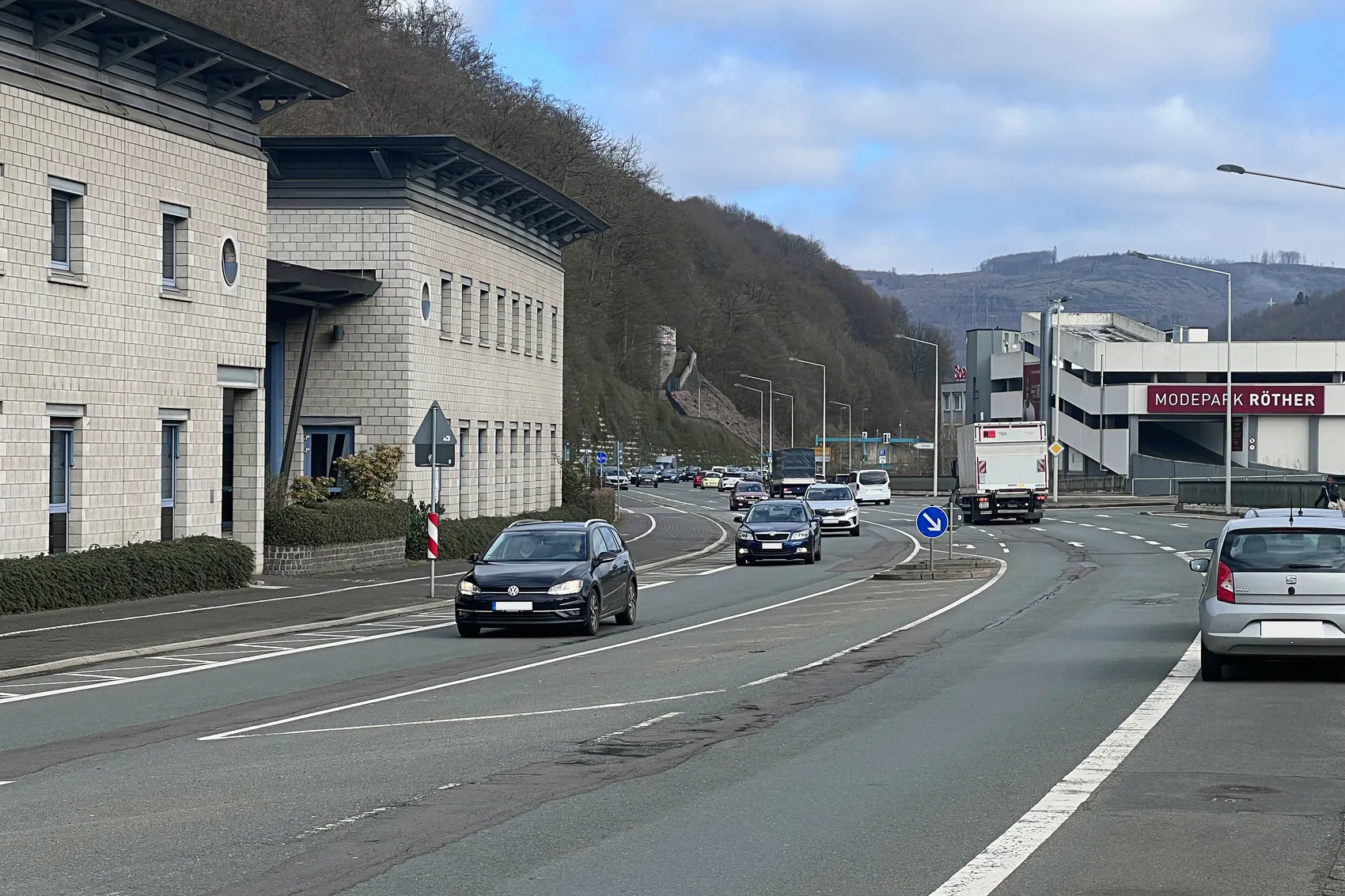Mehrspurige Straße mit fahrenden Autos, links ein modernes Bürogebäude und rechts ein Parkhaus mit dem Schriftzug „ModePark Röther“.