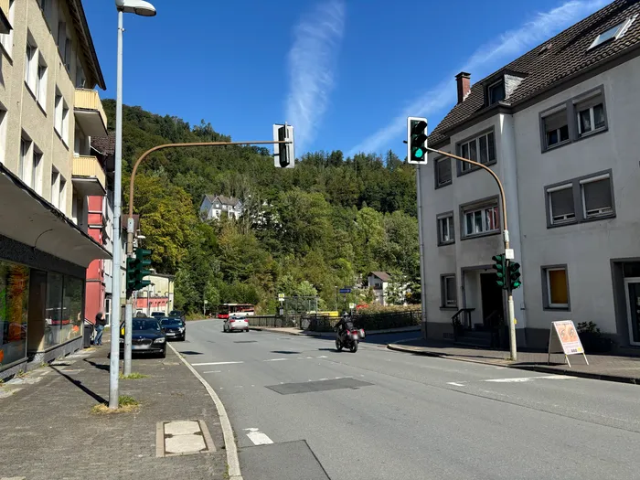 Blick auf eine Straße mit grünen Ampeln, einem Motorradfahrer und Gebäuden vor einem bewaldeten Hang.