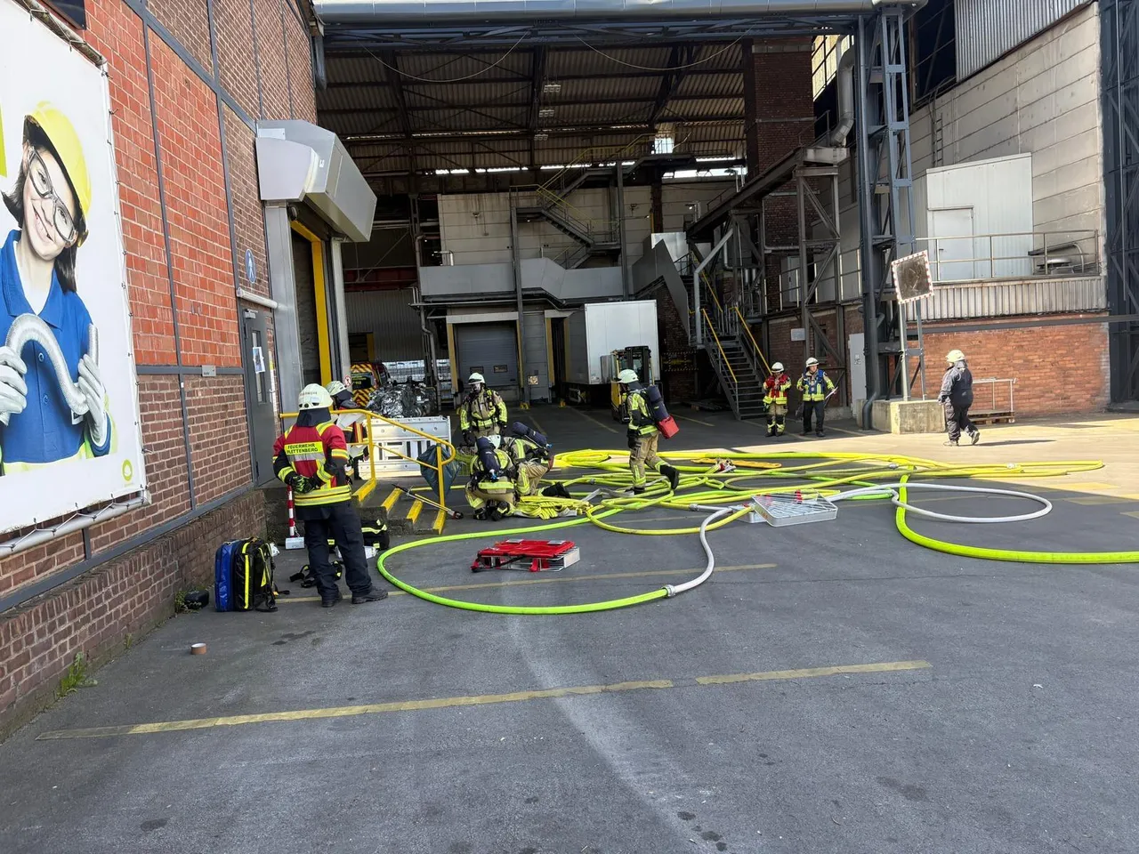 Feuerwehrleute in voller Montur üben auf dem Gelände von Novelis in Ohle, Schläuche liegen bereit. Im Hintergrund eine große Industriehalle.