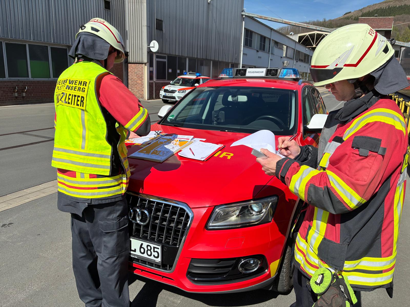 Zwei Feuerwehrleute in voller Montur besprechen Einsatzpläne an der Motorhaube eines roten Autos vor einem Fabrikgebäude.
