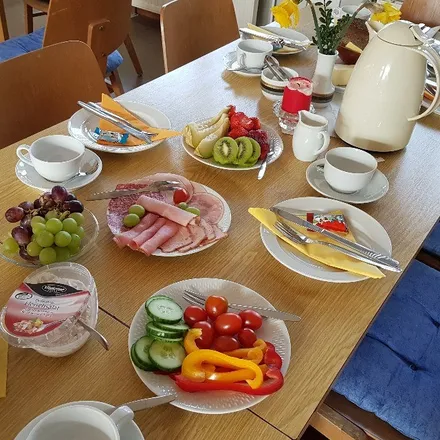 Reich gedeckter Tisch mit verschiedenen Speisen wie Aufschnitt, Obst, Gemüse, Käse und Joghurt sowie Kaffeetassen und einer Kanne.