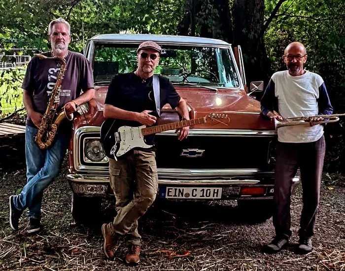 Drei Musiker der Band „Rootz Horns“ posieren mit Saxophon, E-Gitarre und Flöte vor einem braunen Oldtimer-Truck.