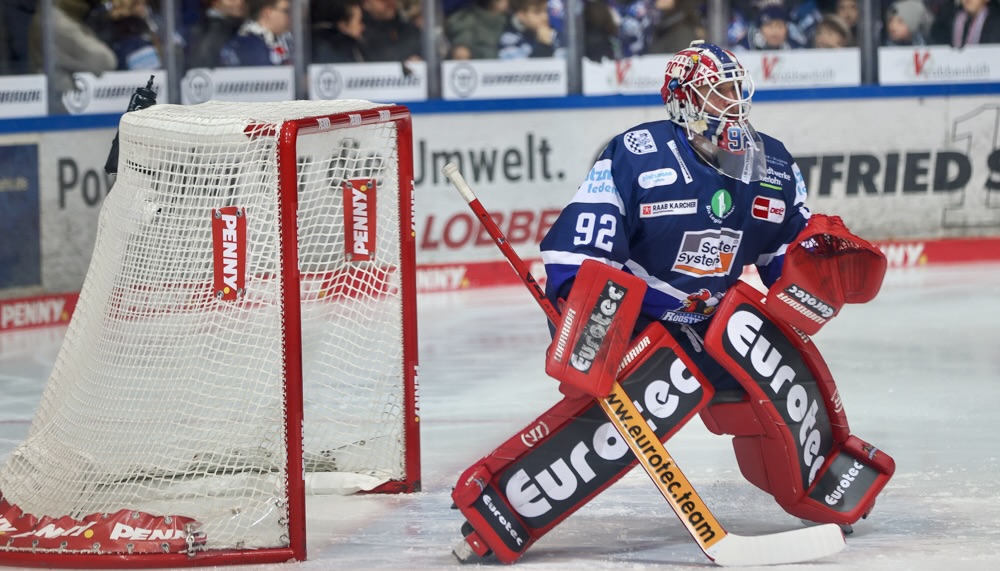 Eishockeytorwart Andreas Jenike in voller Ausrüstung im Tor, bereit für die Abwehr. Sein Trikot trägt die Nummer 92 und verschiedene Werbelogos.