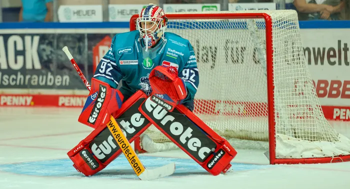 Eishockey-Torwart Andreas Jenike von den Iserlohn Roosters in blauem Trikot und roten Pads steht vor dem Tor.