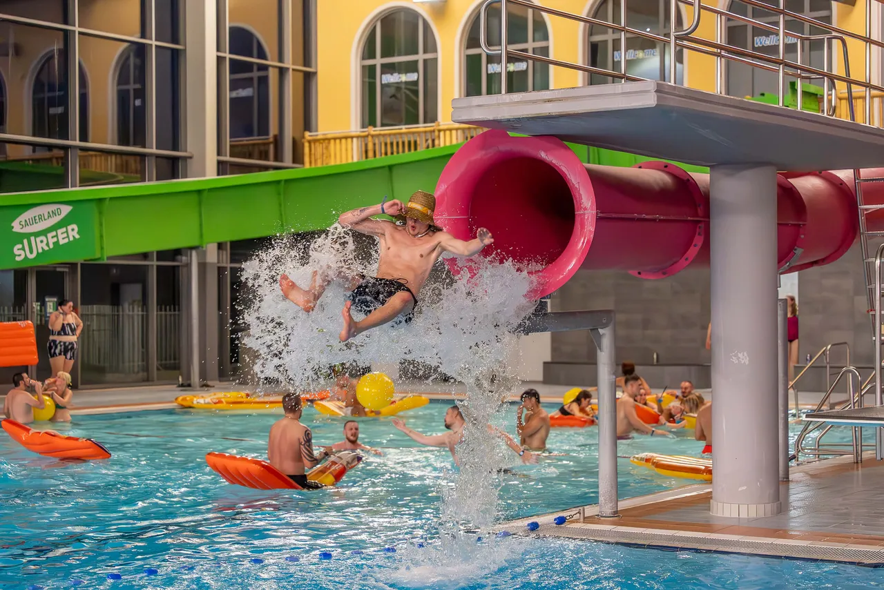 Ein Mann springt aus einer rosa Wasserrutsche in ein Schwimmbecken und erzeugt eine große Wasserfontäne.