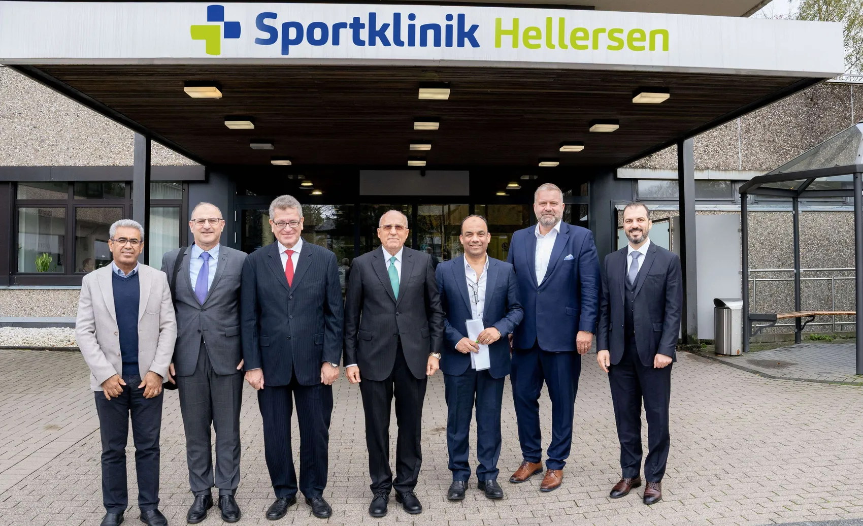 Sieben Männer in Anzügen stehen vor dem Eingang der Sportklinik Hellersen. Im Hintergrund ist das Logo der Klinik zu sehen.