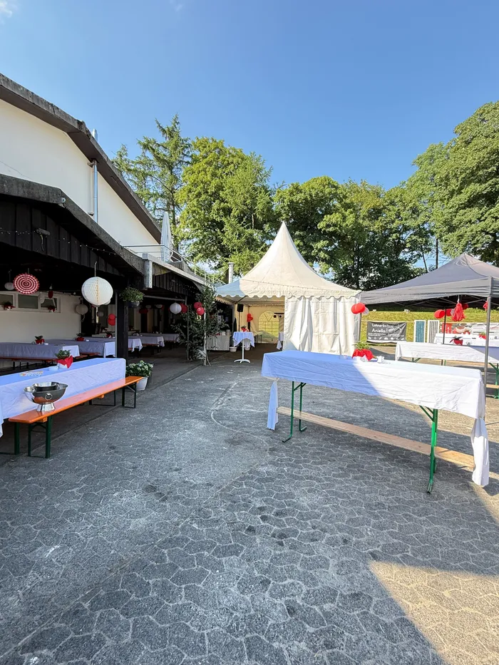 Zwei Festzelte stehen auf einem gepflasterten Hof vor einem traditionellen Fachwerkhaus, dekoriert mit roten Lampions. Tische mit weißen Tischdecken stehen bereit, ein Bierwagen ist zu sehen.