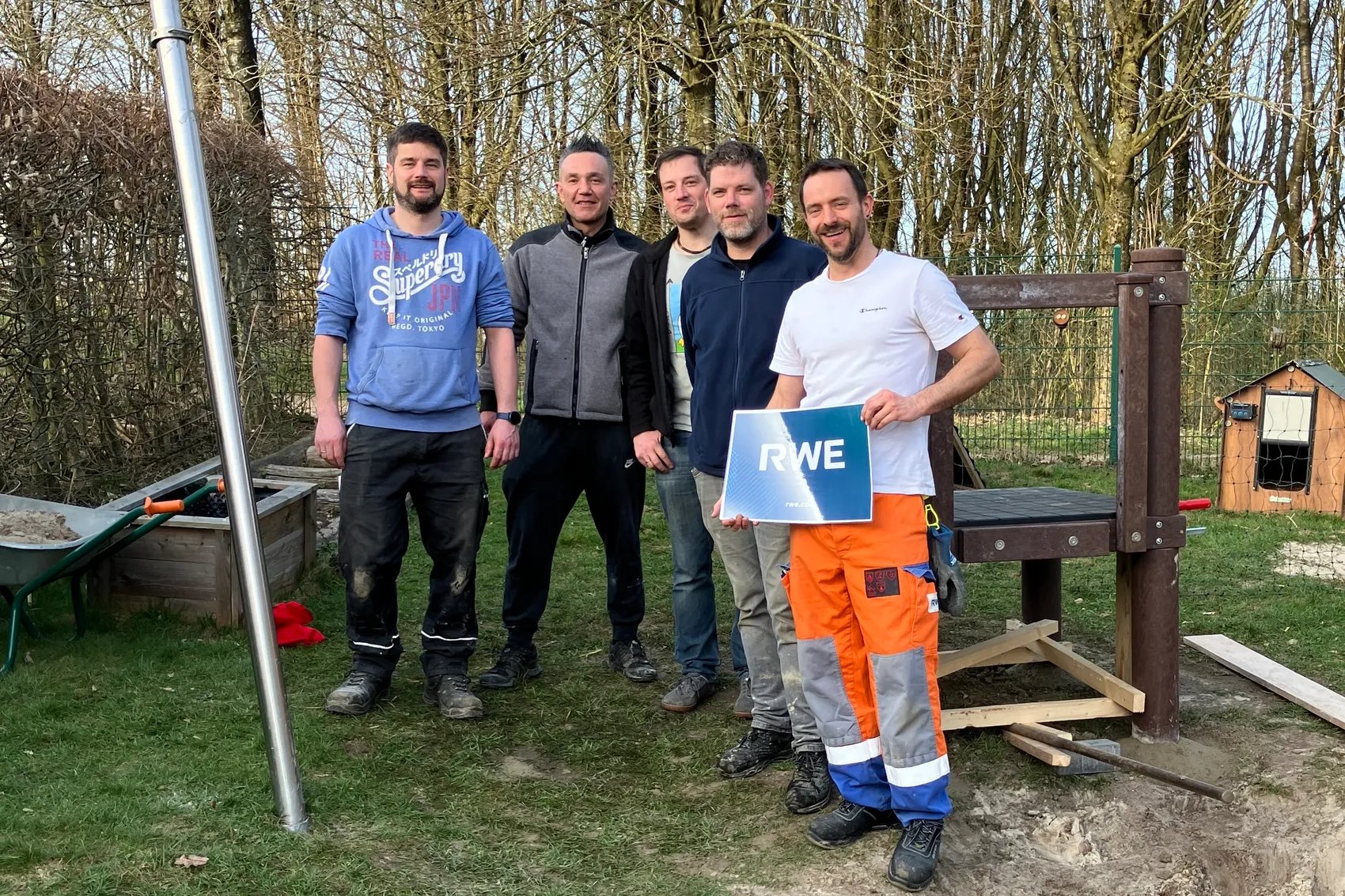 Fünf Männer stehen lächelnd vor einer fast fertig montierten Rutsche auf dem Gelände einer Kindertagesstätte. Ein Mann hält ein Schild mit dem RWE-Logo.