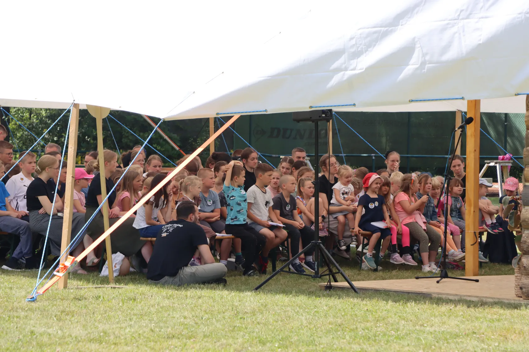 Eine Gruppe Kinder sitzt auf Bänken unter einem weißen Zelt und schaut auf eine Person, die vor ihnen kniet. Im Hintergrund ist ein Banner mit dem Schriftzug „Duni“ zu sehen.