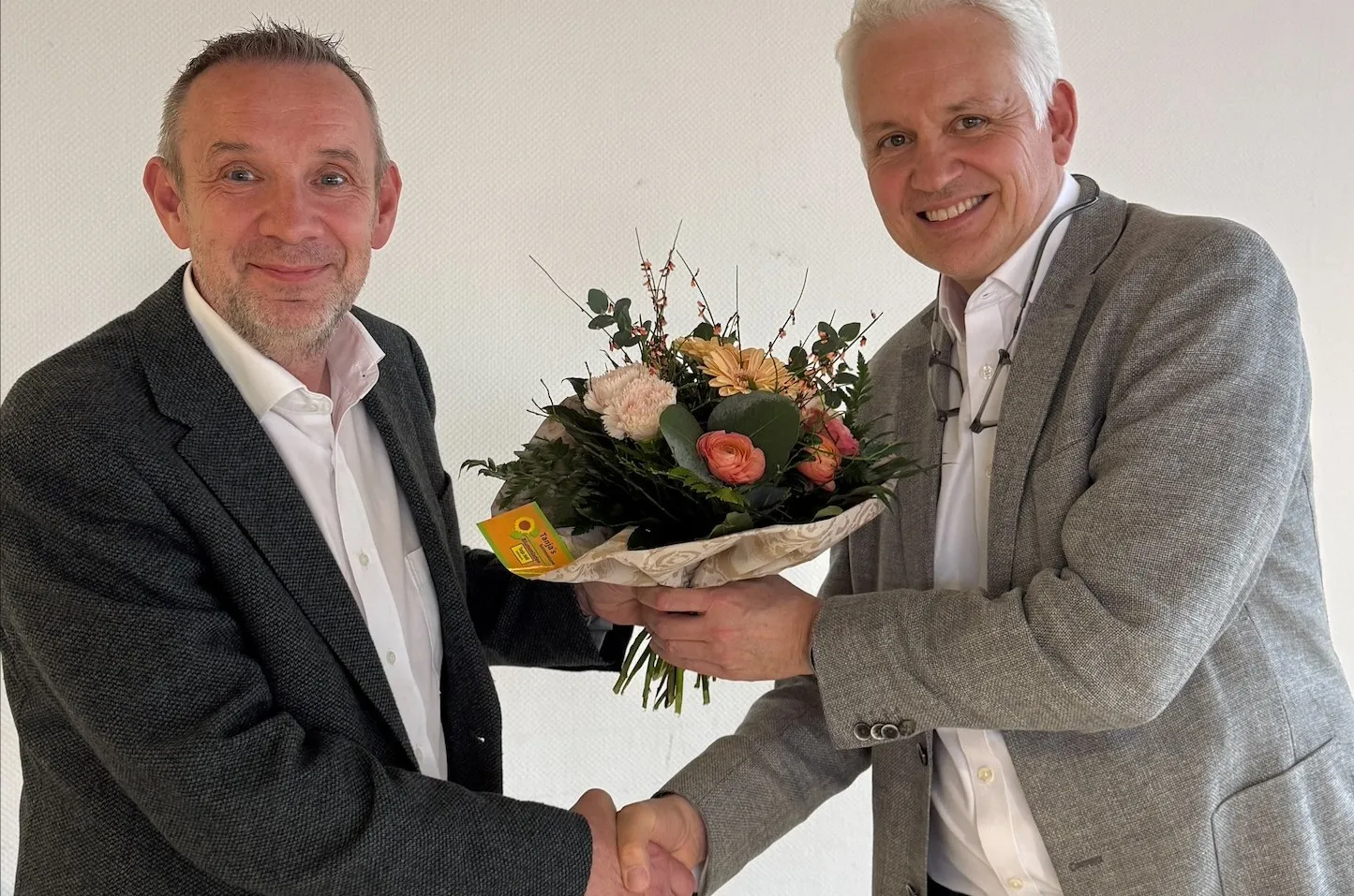 Zwei Männer im Anzug geben sich die Hände, während einer von ihnen einen Blumenstrauß hält. Im Hintergrund eine weiße Wand.