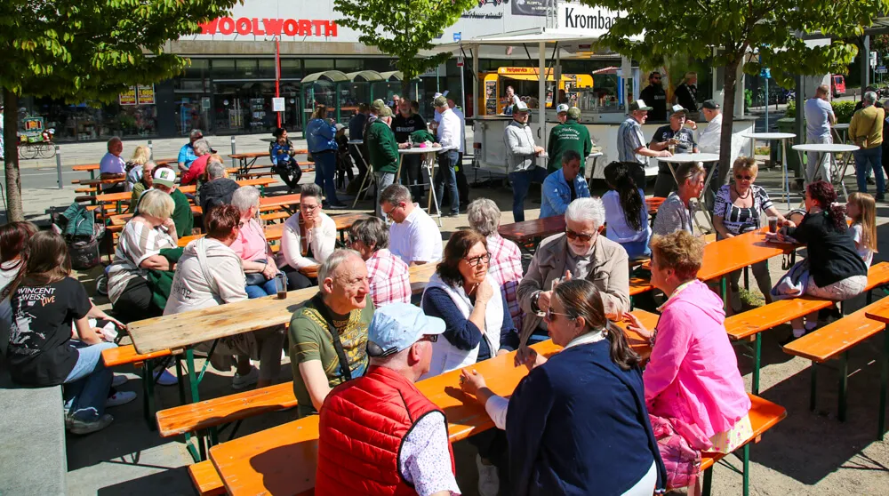 Menschen sitzen und stehen an Biertischgarnituren auf einem Platz, im Hintergrund Geschäfte und ein Imbissstand. Viele Besucher sind beim Maifest in Letmathe versammelt.