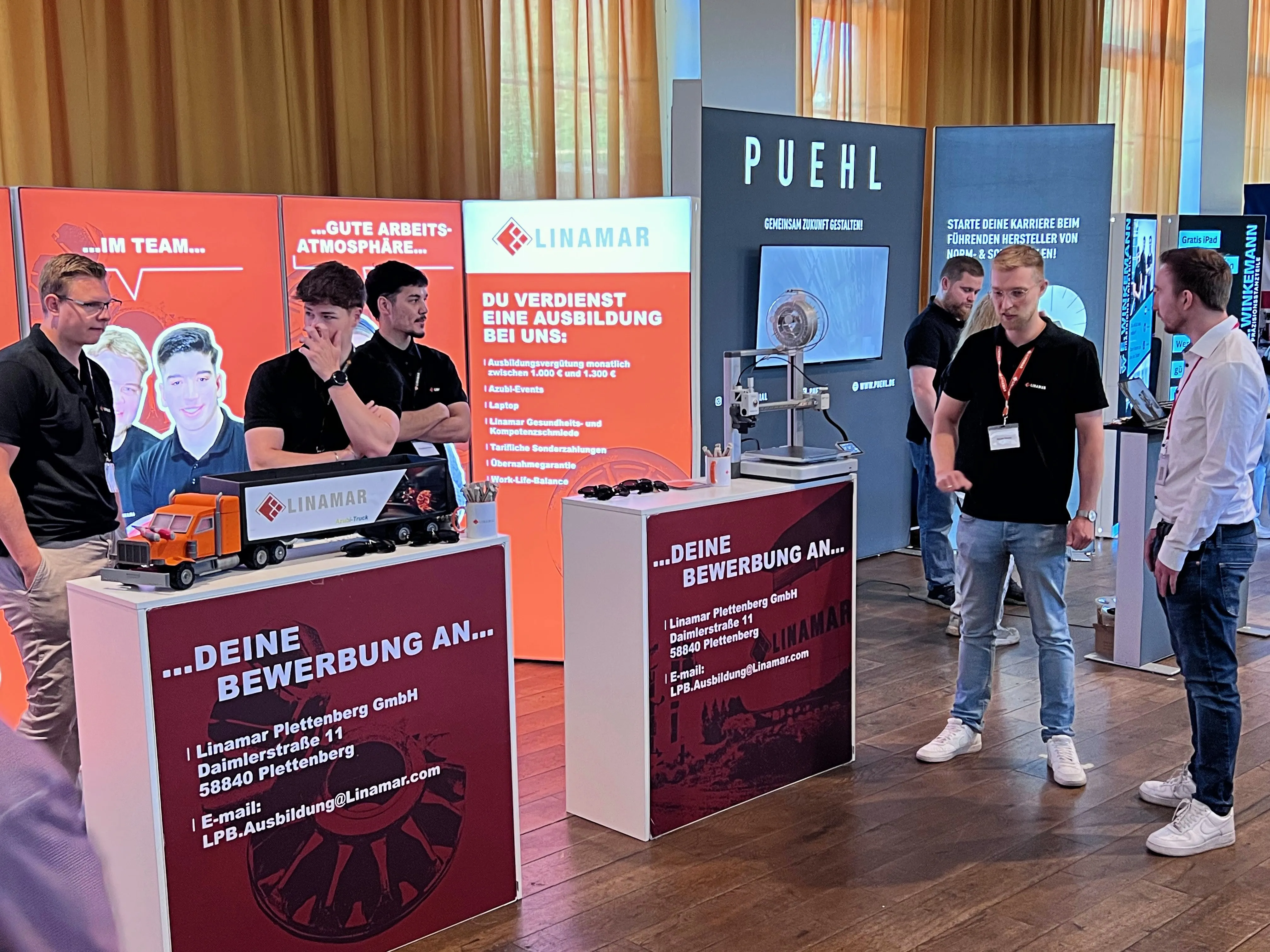 Messestand von Linamar mit Informationen zu Ausbildungsplätzen, Mitarbeitern und Fahrzeugen. Im Hintergrund weitere Stände und Besucher einer Jobmesse.