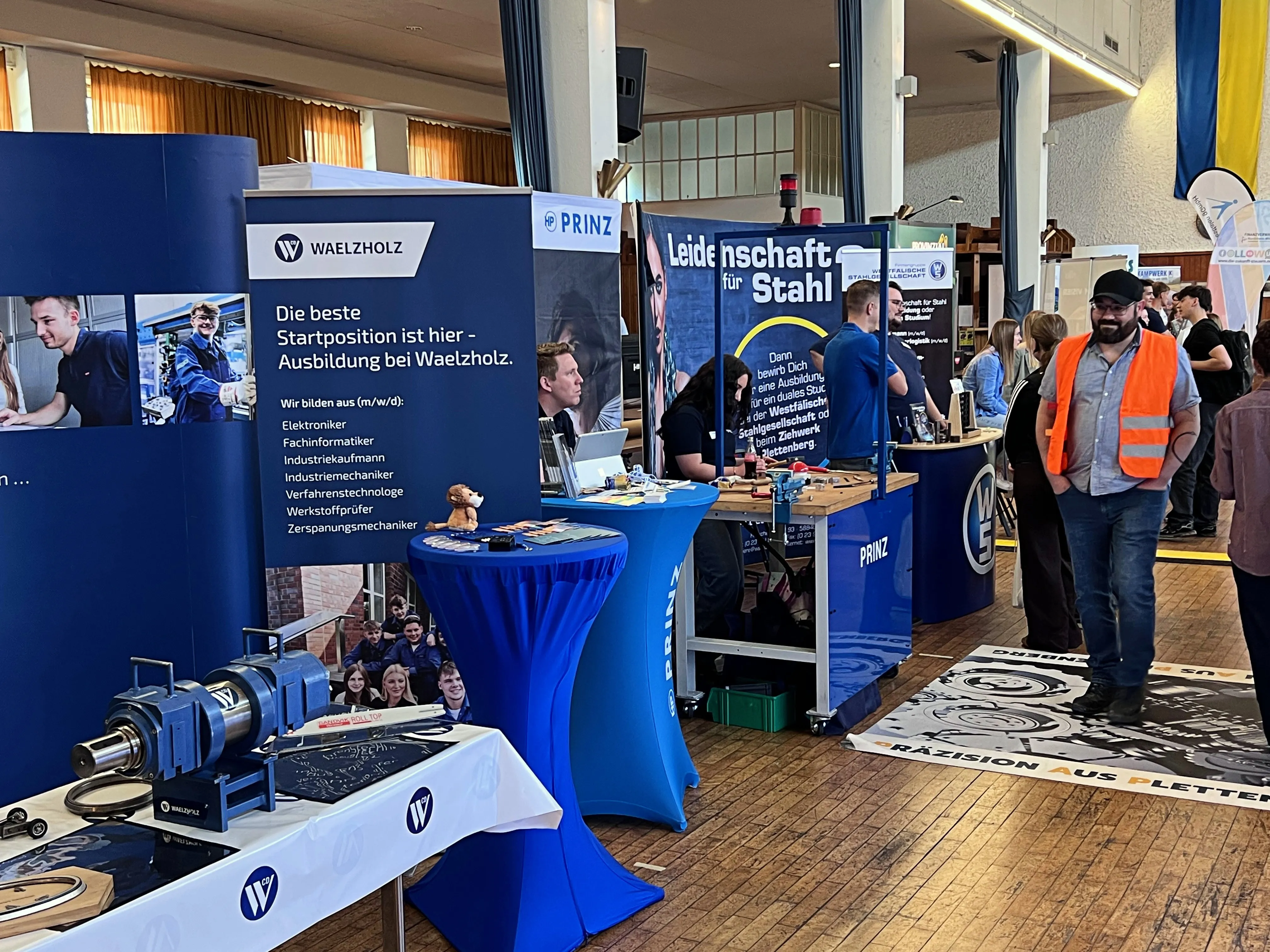 Zwei Beratende von Waelzholz und Prinz sitzen an einem Messestand und sprechen mit einem jungen Mann. Im Hintergrund sind Werbebanner und weitere Messestände zu sehen.