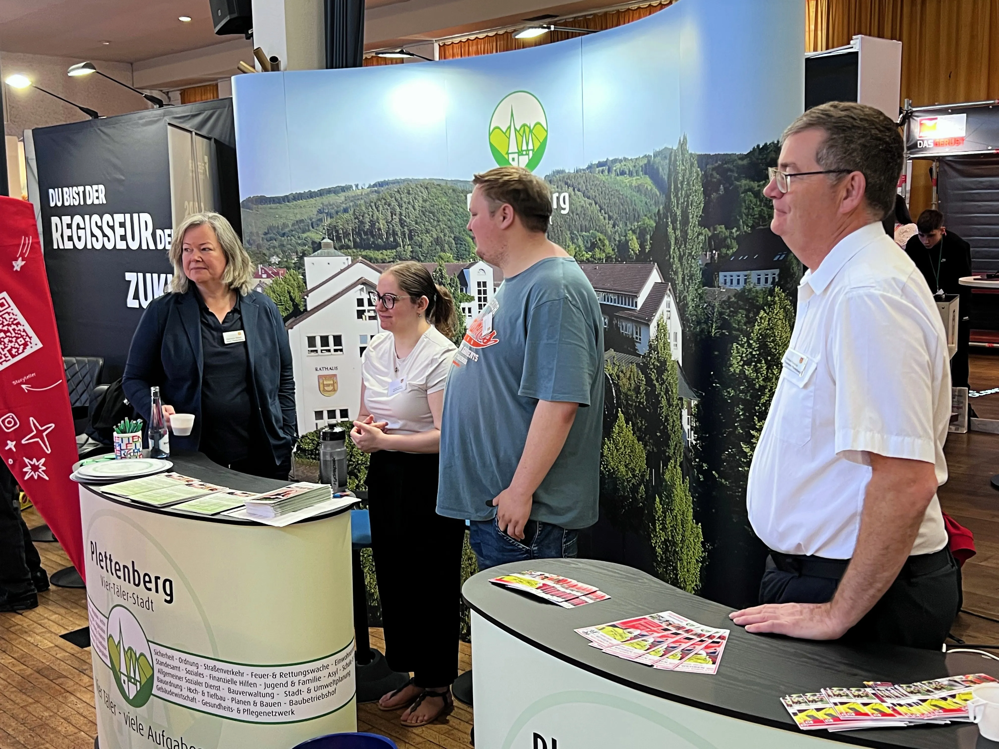 Vier Personen stehen an einem Messestand mit dem Logo der Stadt Plettenberg vor einer Kulisse mit Wald und Hügeln. Sie verteilen Informationsmaterial und beraten Interessierte auf der Ausbildungsbörse.
