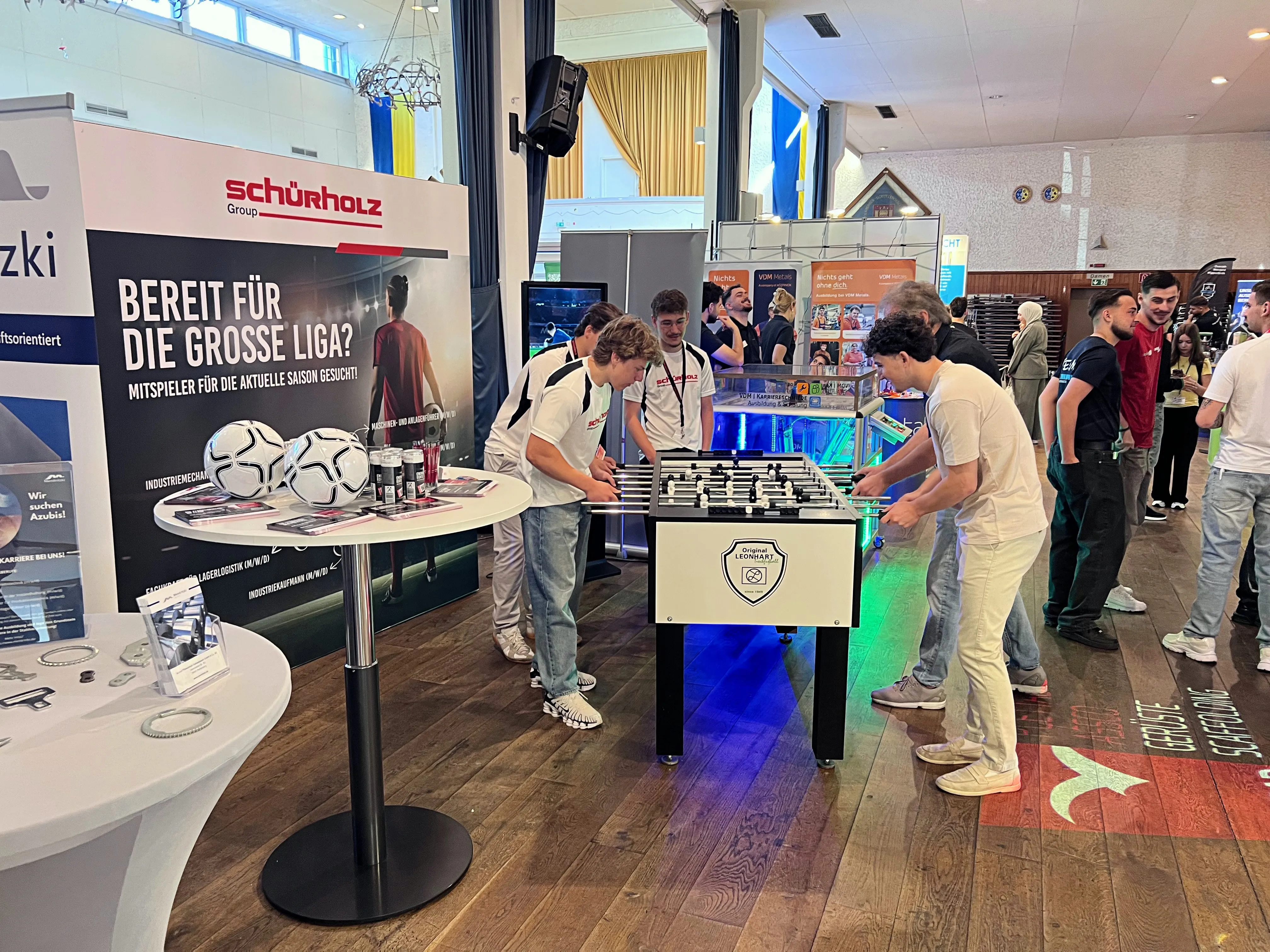 Zwei junge Männer spielen auf einem beleuchteten Tischfußballspiel vor einem Stand mit dem Schriftzug „Schürholz Group“ und einem großen Fußball. Im Hintergrund stehen weitere Besucher einer Ausbildungsbörse in einer Halle.