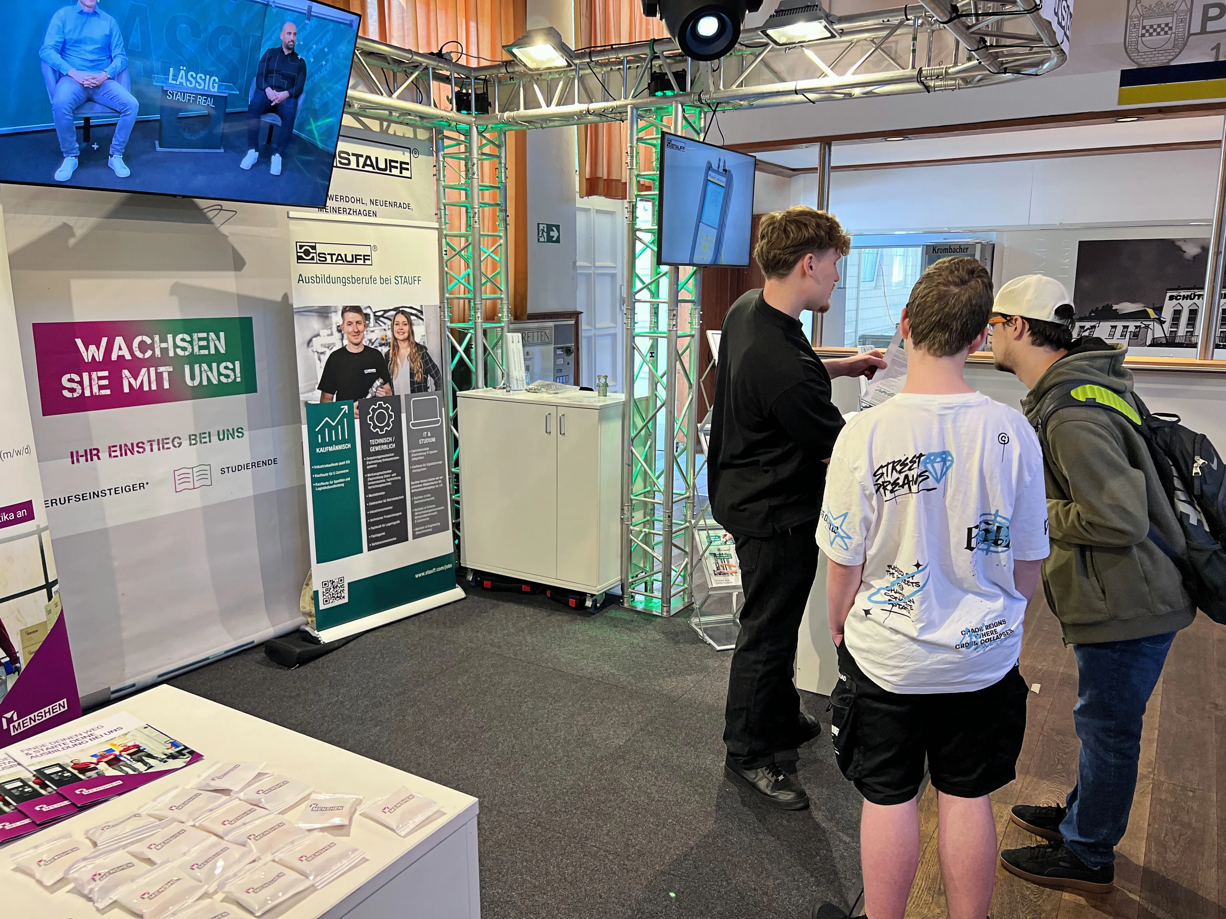 Zwei Jugendliche betrachten einen Messestand von STAUFFE, der Ausbildungsangebote bewirbt. Im Hintergrund sind weitere Besucher und ein Vortragender auf einer Bühne zu sehen.