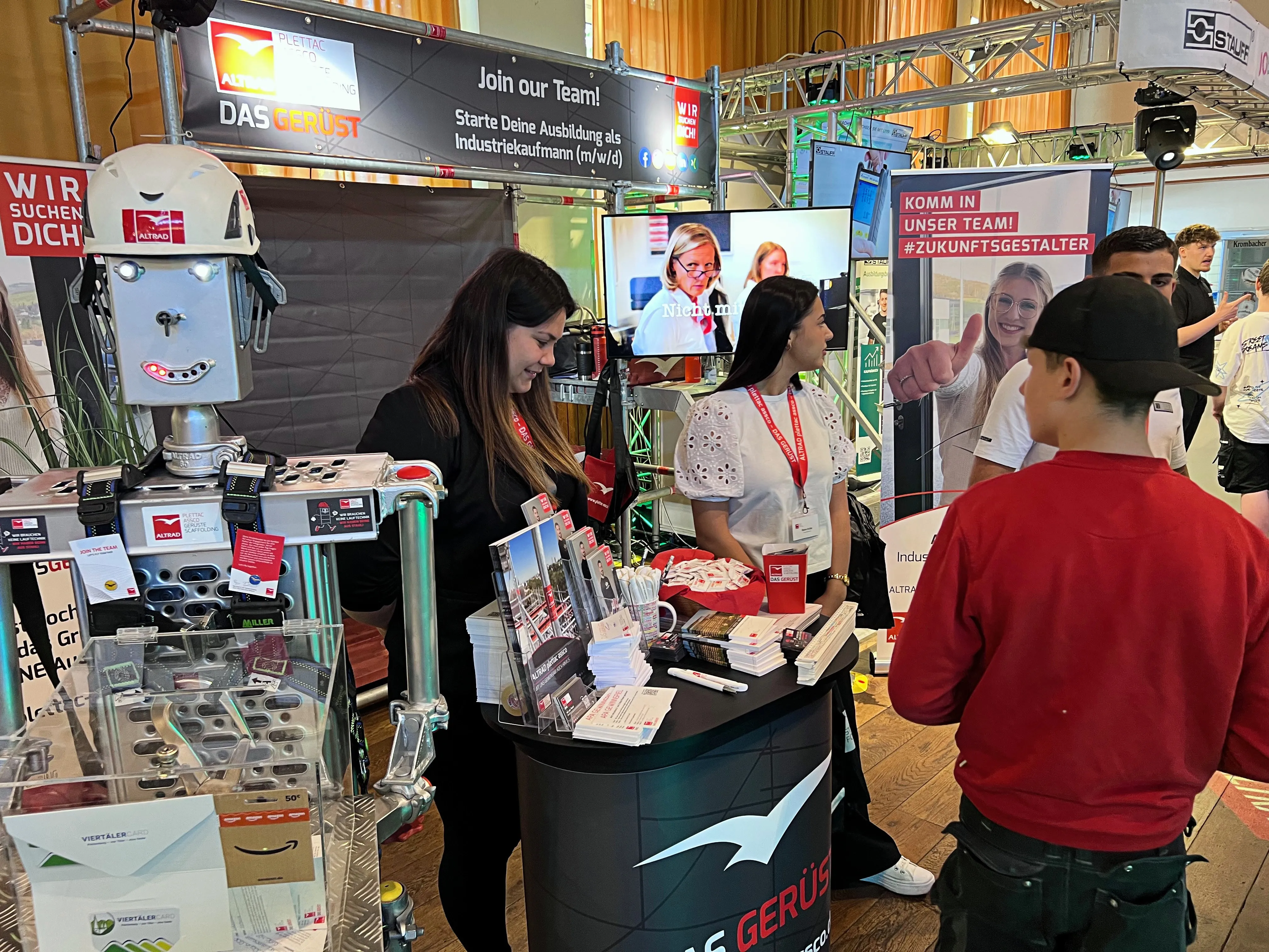 Zwei Mitarbeiterinnen eines Unternehmens stehen an einem Messestand mit Informationsmaterialien, während ein junger Mann mit rotem Polohemd und ein weiterer Jugendlicher sich informieren.