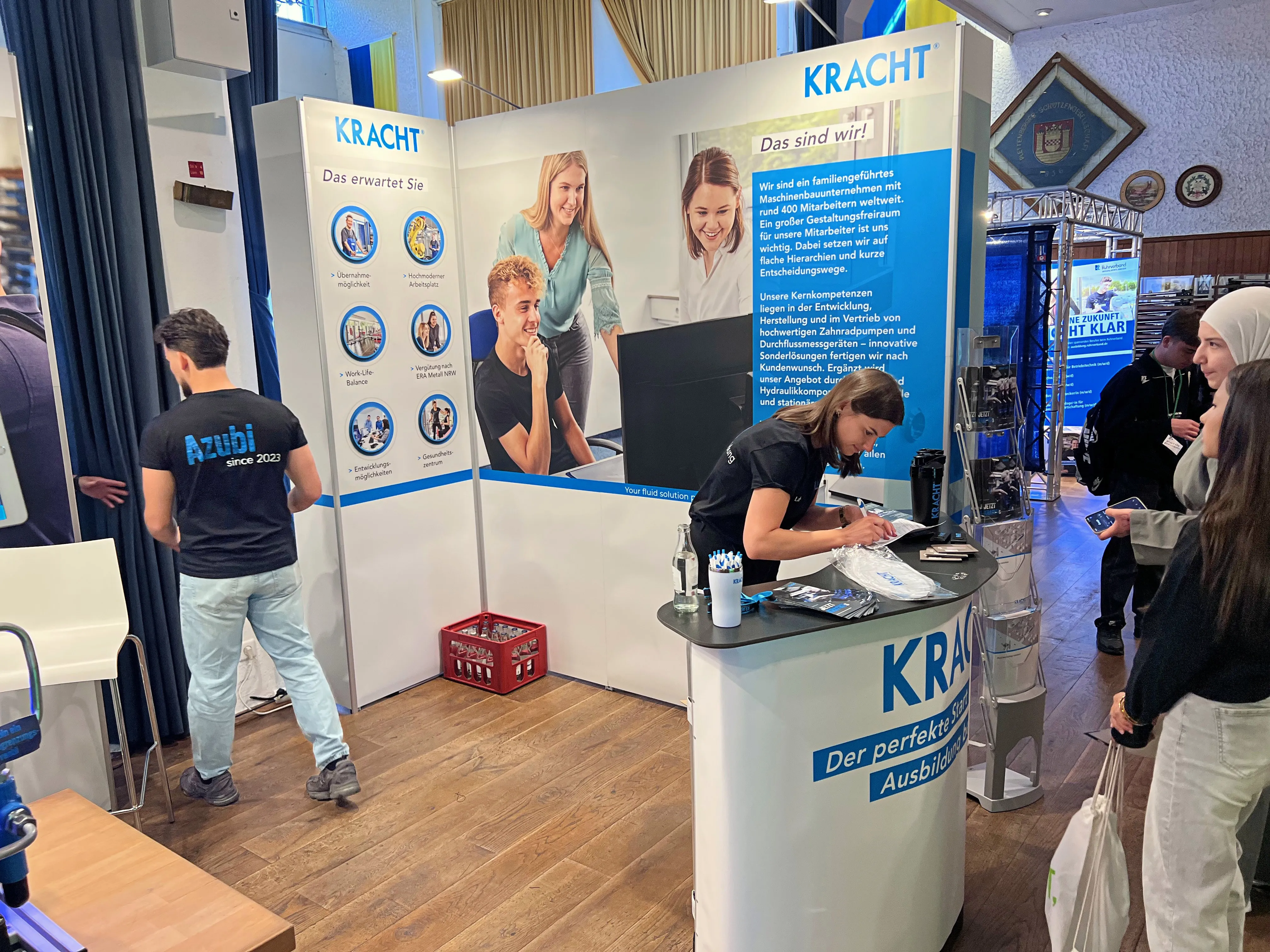Zwei junge Männer stehen an einem Messestand mit dem Logo von „KRACHT“ und Informationsmaterialien über Ausbildungsangebote. Im Hintergrund sind weitere Stände und Besucher einer Ausbildungsbörse zu sehen.