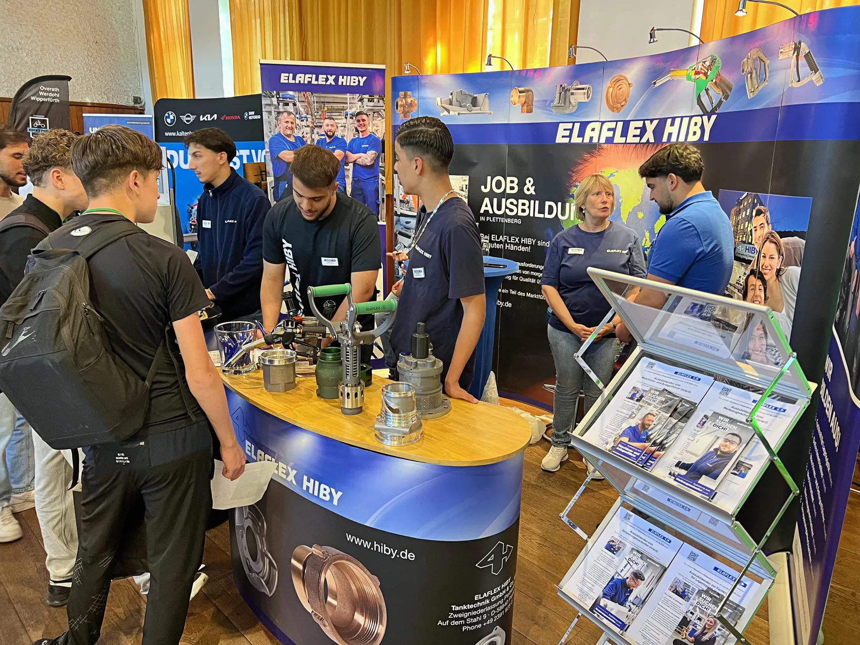 Mehrere junge Männer betrachten an einem Messestand verschiedene Metallteile und Werkzeuge, während ein Mitarbeiter sie berät. Im Hintergrund sind Werbebanner für ELAFLEX HIBY zu sehen.