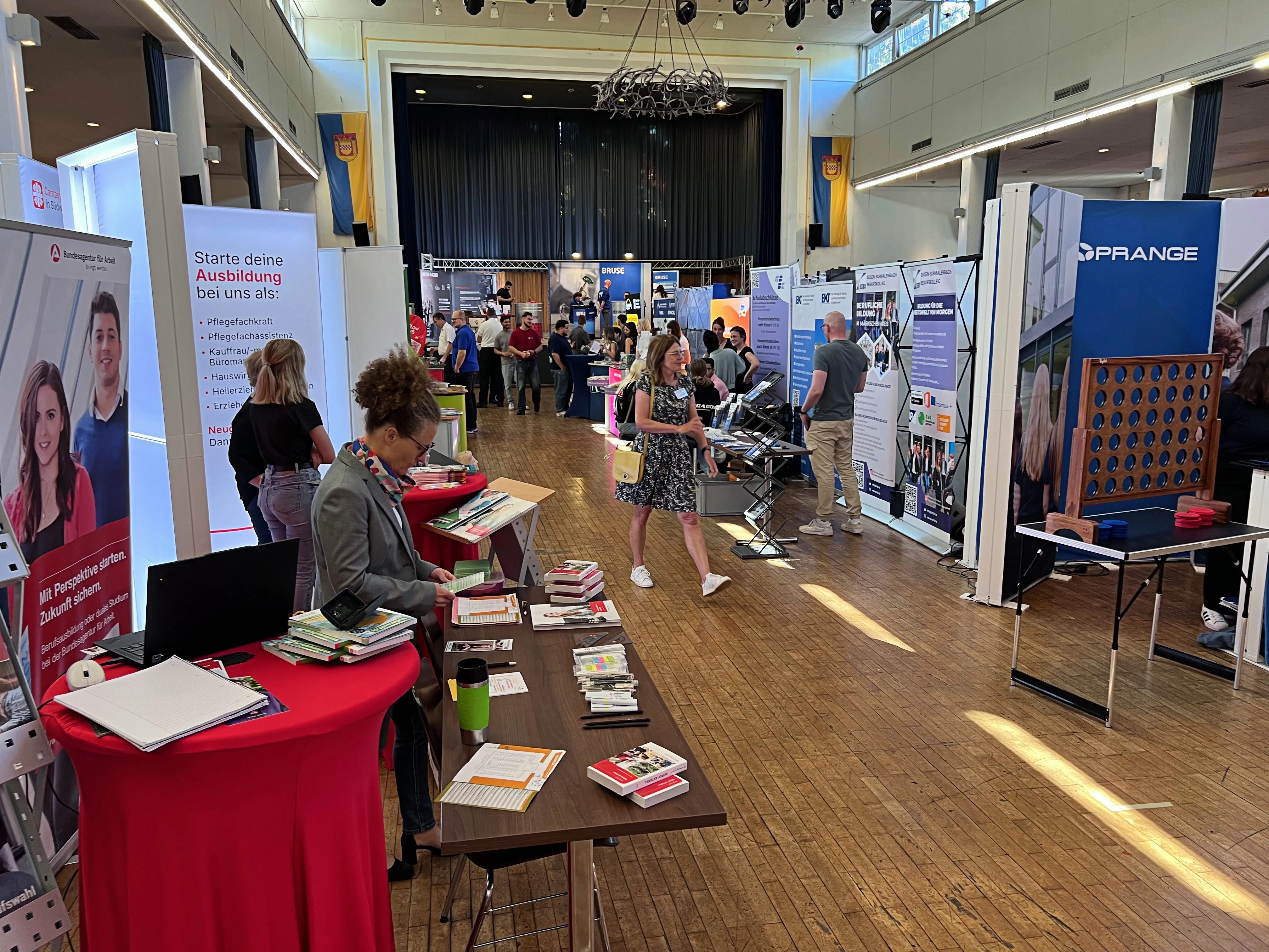 Mehrere Stände mit Informationsmaterialien und Ansprechpartnern auf einer Ausbildungsbörse in einer großen Halle. Im Vordergrund ein Stand mit roten Tischdecken und Broschüren, dahinter weitere Stände mit Roll-ups und Firmenschildern.