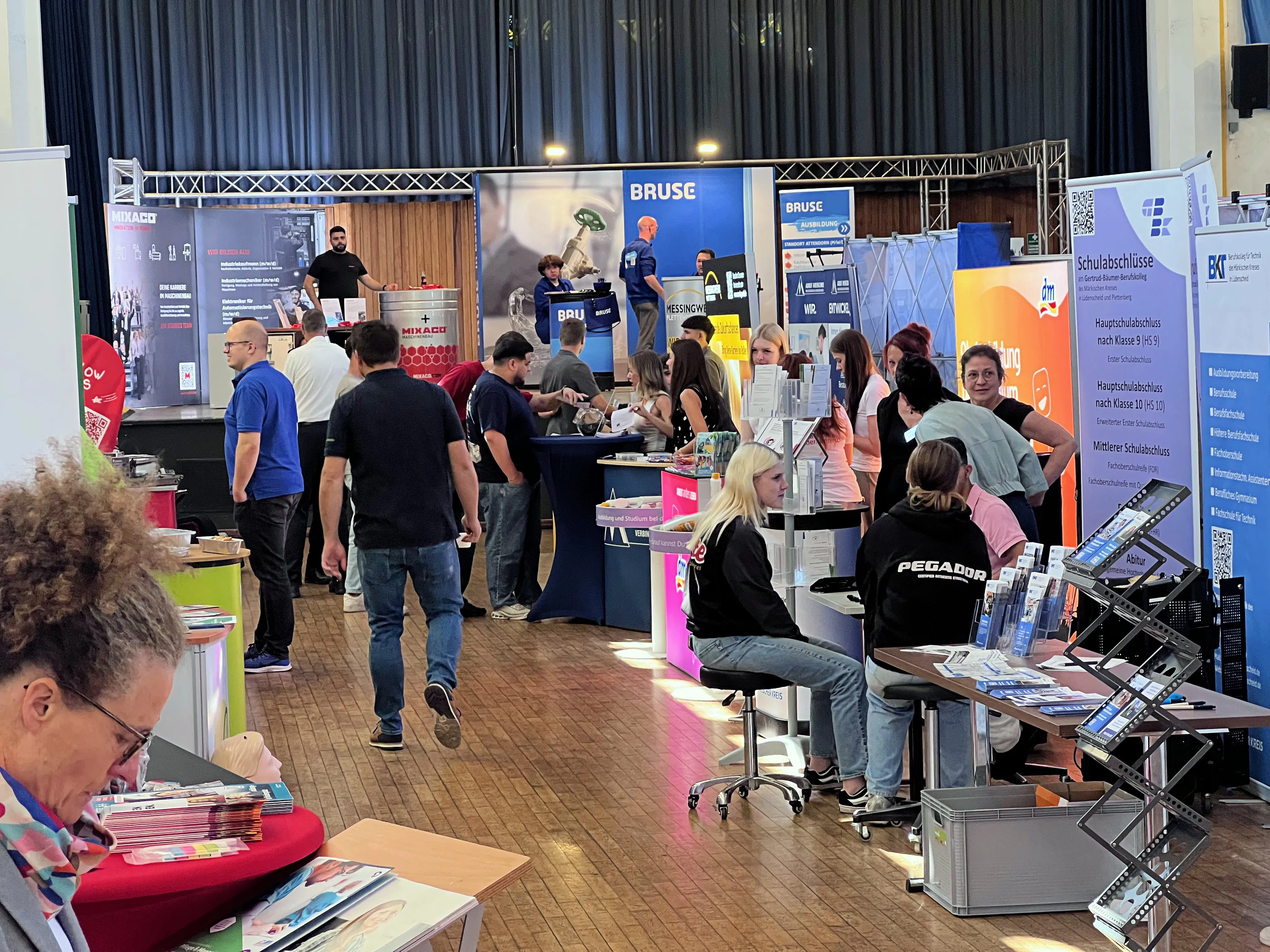 Belebte Messehalle mit Ständen von Unternehmen und Organisationen, die über Ausbildungsangebote informieren. Besucher stehen an den Ständen und unterhalten sich mit den Ausstellern.