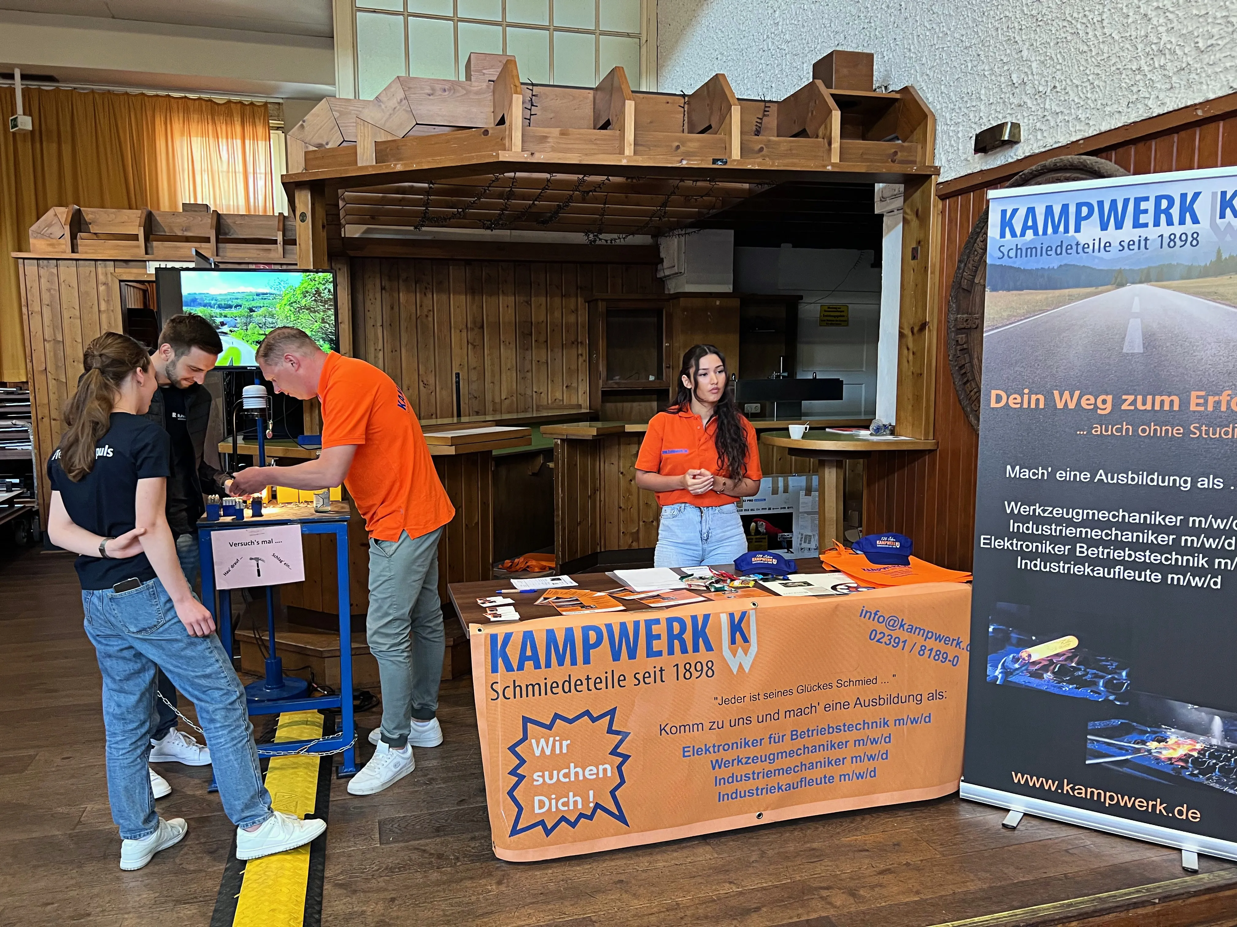 Drei junge Männer und eine junge Frau stehen an einem Messestand von Kampwerk Schmiedeteile, der Ausbildungsplätze anbietet. Im Hintergrund ist die belebte Ausbildungsbörse in der Plettenberger Schützenhalle zu sehen.
