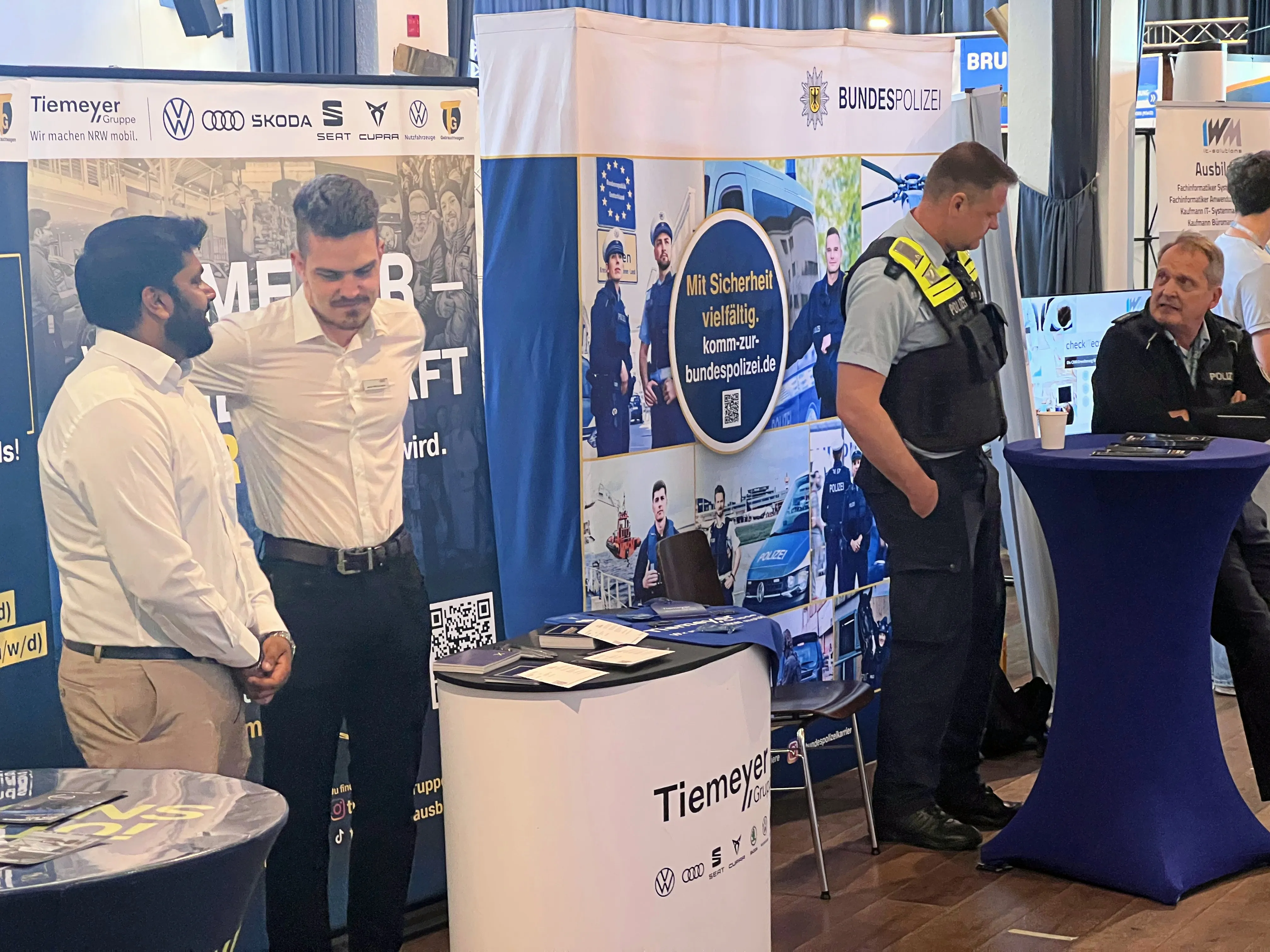 Ein Polizist steht an einem Infostand der Bundespolizei auf einer Ausbildungsbörse. Im Hintergrund ein Banner mit Bildern von Polizisten und dem Slogan „Mit Sicherheit vielfältig. komm.zur.bundespolizei.de“.