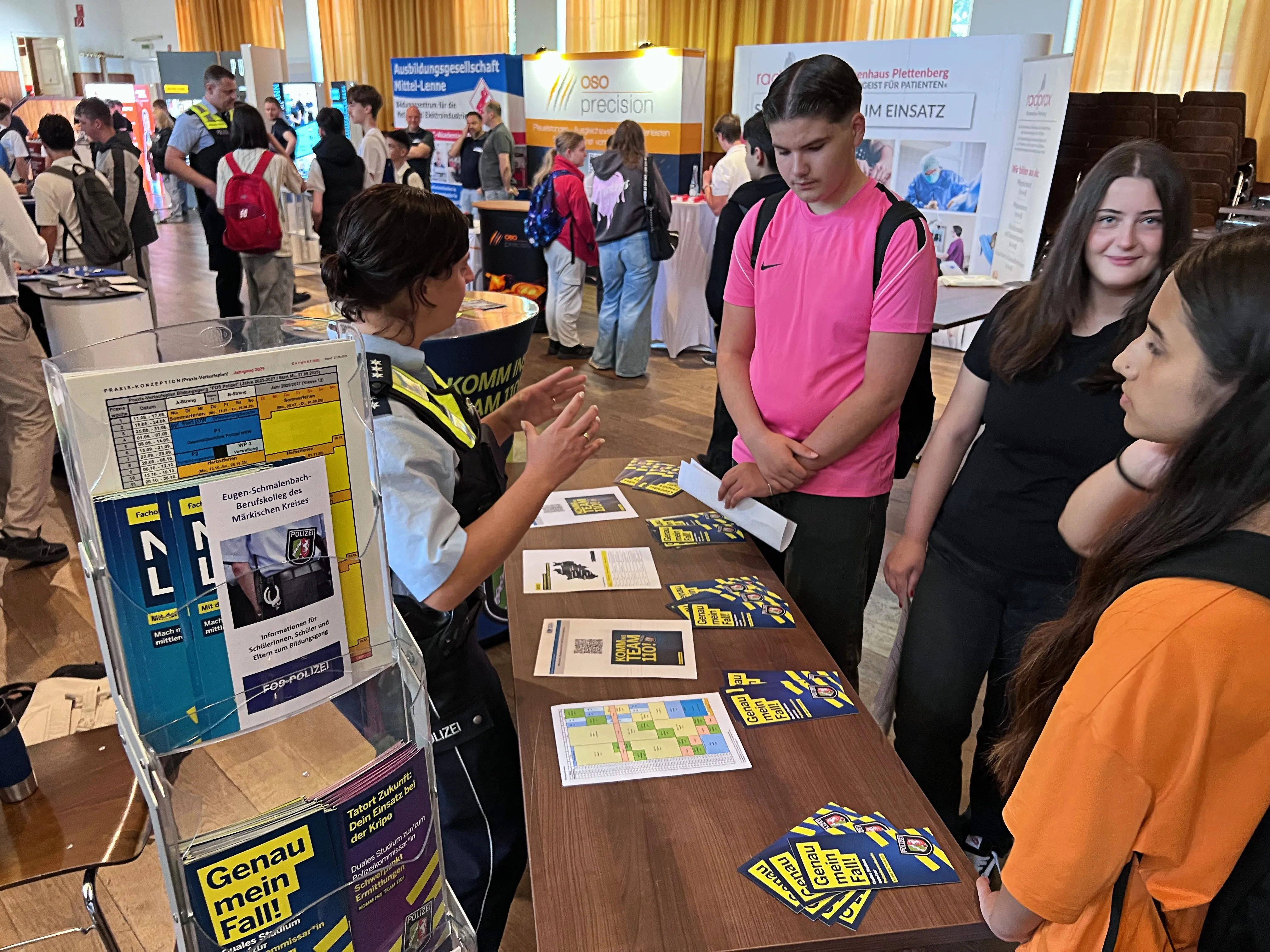 Zwei junge Frauen an einem Messestand mit Informationsmaterialien und einem Spiel zur Berufsorientierung interagieren mit einer Person in Uniform. Im Hintergrund sind weitere Besucher und Stände auf einer Ausbildungsbörse zu sehen.