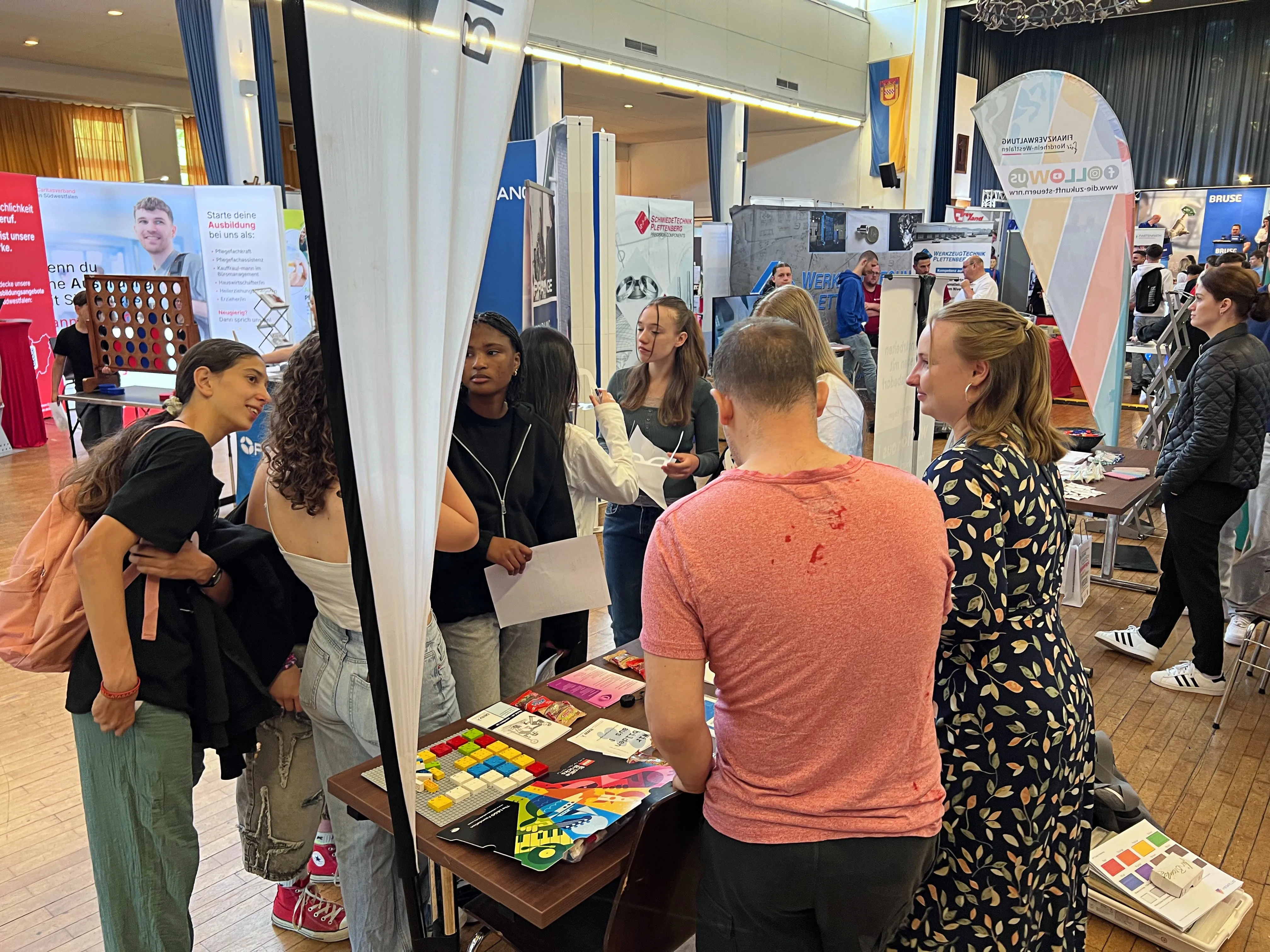 Mehrere junge Menschen stehen an einem Messestand und sprechen mit einer Berufsberaterin. Im Hintergrund sind weitere Stände und Besucher einer Ausbildungsbörse zu sehen.