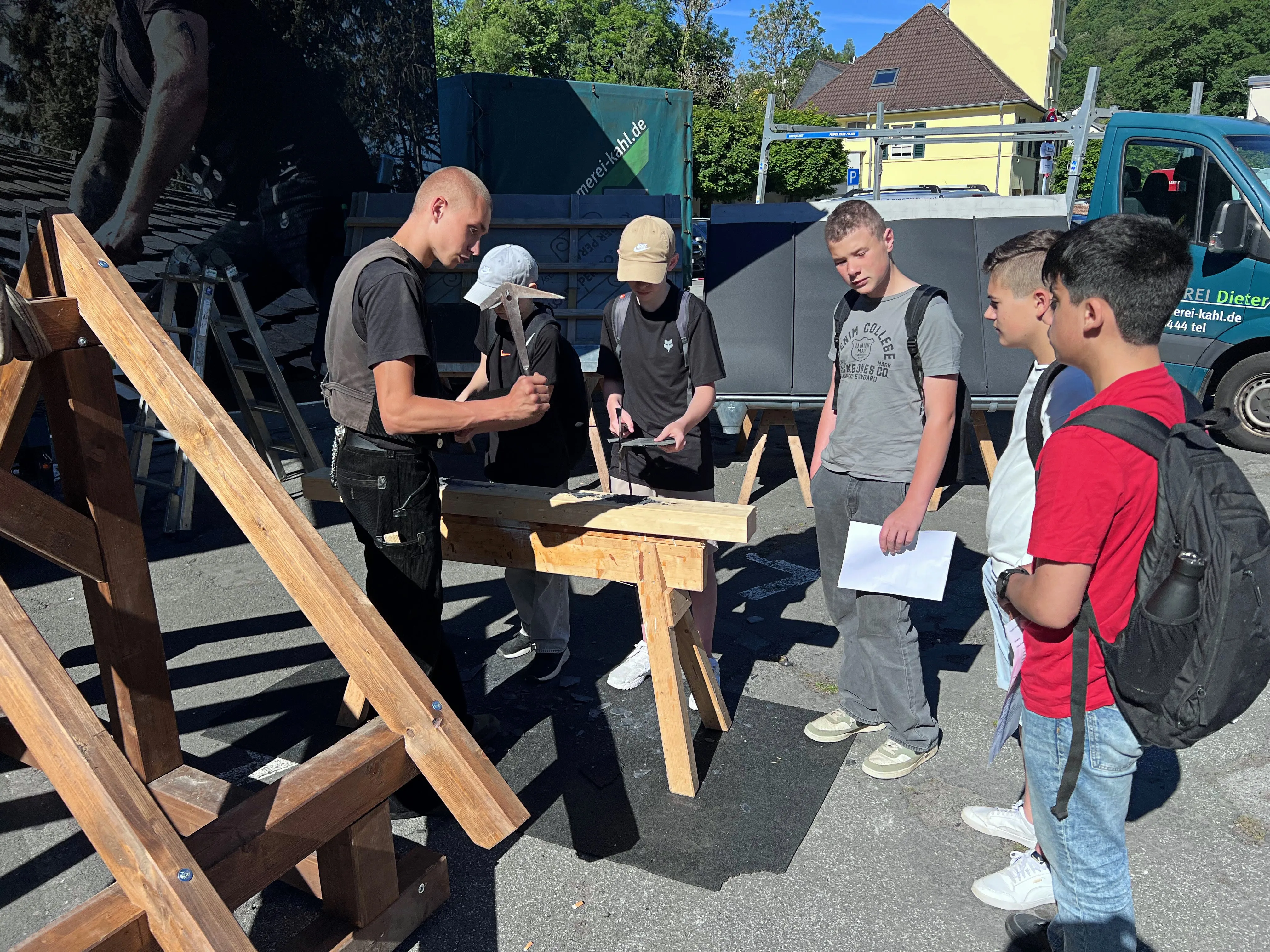 Ein Ausbilder erklärt Jugendlichen an einem Holztisch im Freien die Handhabung von Werkzeugen. Im Hintergrund ein Transporter und weitere Personen.