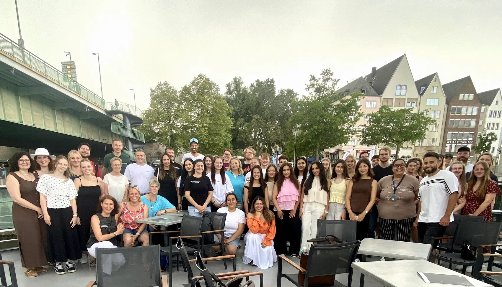 Eine große Gruppe von Menschen posiert für ein Gruppenfoto auf einer Außenterrasse vor Gebäuden und einer Brücke in Köln.