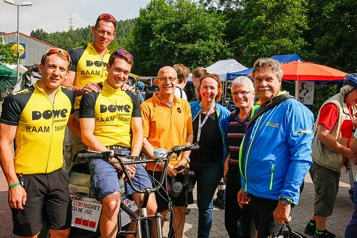 Eine Gruppe von Menschen, darunter Radfahrer in gelben Trikots mit der Aufschrift „DOWE RAAM team“, posiert gemeinsam mit Fahrrädern im Freien.