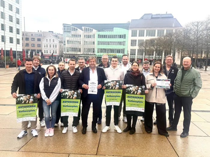 Gruppe von etwa 20 Personen, hauptsächlich Männer in Anzügen, posieren mit Flyern für die 15. Autoschau auf dem Rathausplatz in Lüdenscheid.