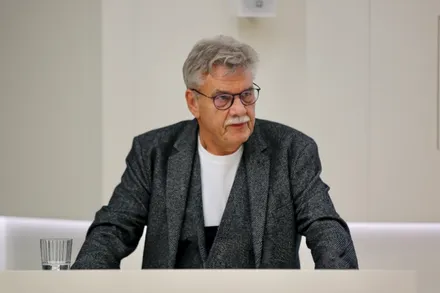 Ein Mann mit grauen Haaren und Schnurrbart sitzt am Tisch und blickt zur Seite. Er trägt eine Brille und einen grauen Anzug über einem weißen Hemd. Im Hintergrund eine weiße Wand und eine Deckenmontage.