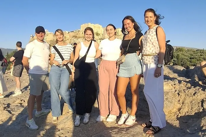 Eine Gruppe von sechs Personen steht lächelnd auf felsigem Gelände vor einer antiken Tempelanlage unter blauem Himmel.