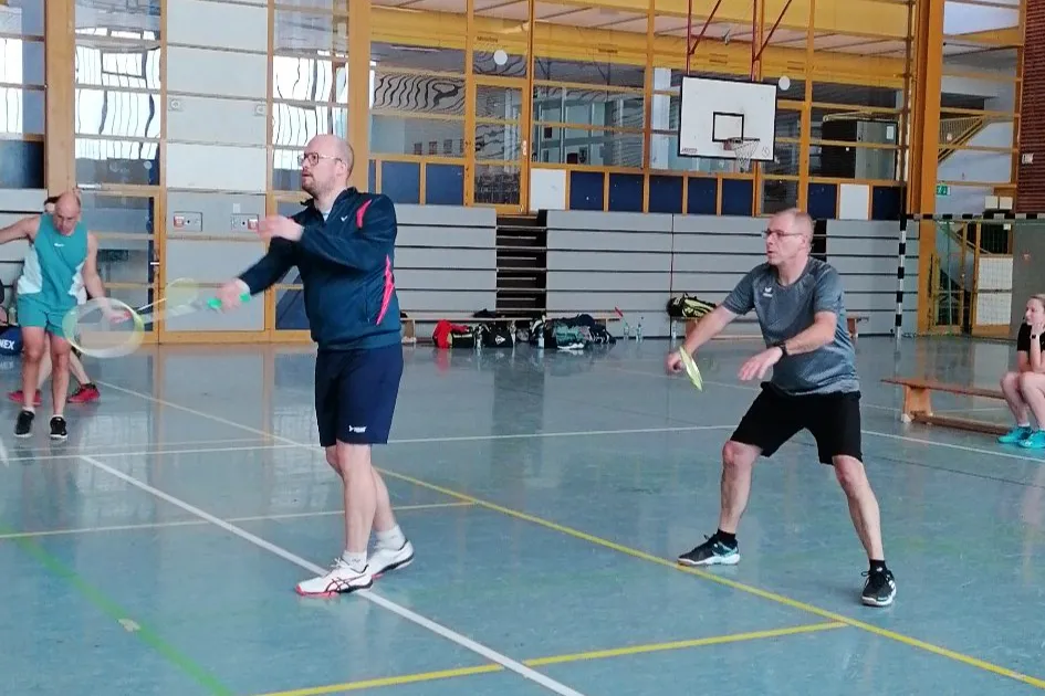 Zwei Badmintonspieler im Spiel, der linke schlägt zu, der rechte bereitet sich auf den Return vor. Im Hintergrund weitere Spieler in einer Sporthalle.
