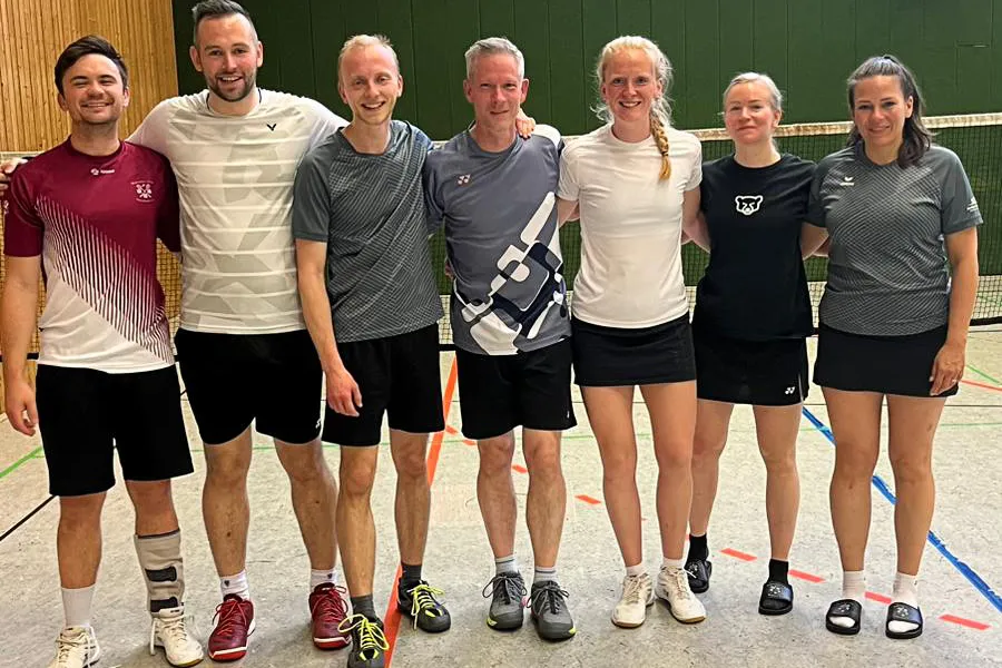 Sechs Badmintonspieler des BC Herscheid stehen lächelnd in Sportkleidung in einer Sporthalle. Im Hintergrund sind Badmintonfelder erkennbar.