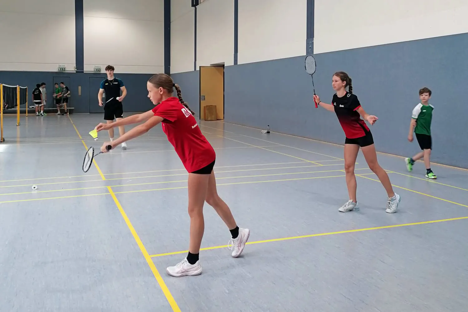 Zwei junge Badmintonspielerinnen schlagen in einer Sporthalle einen gelben Shuttlecock. Im Hintergrund sind weitere Spieler und ein Netz zu sehen.